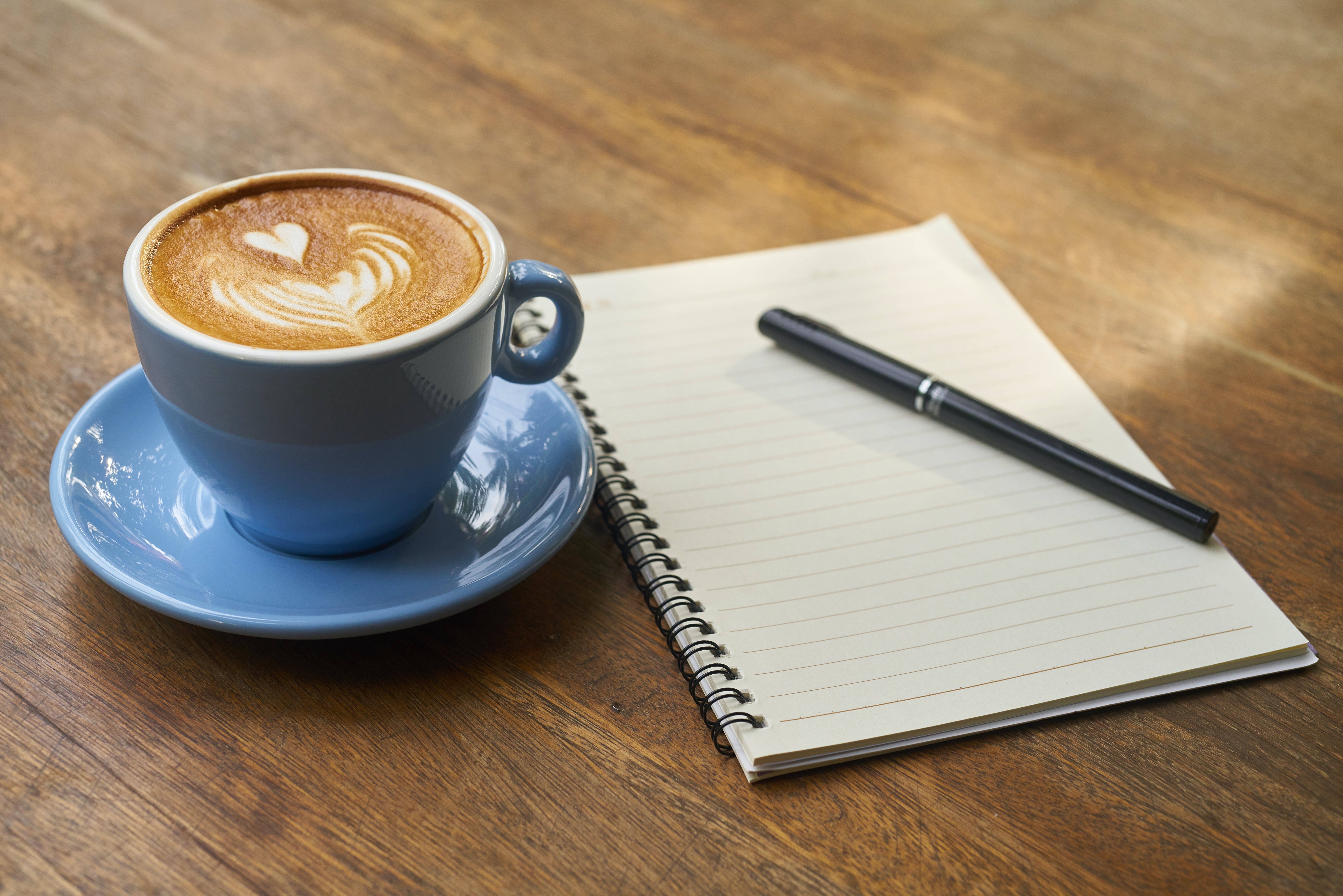 An image of a notebook with a pen on the top with a coffee next to it